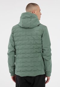 Mann in einer salbeifarbigen, gesteppte Jacke mit Kapuze, der mit dem Rücken zu einem schlichten weißen Hintergrund steht.