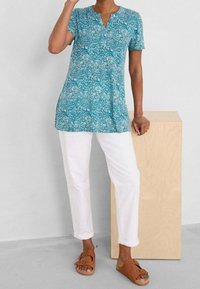 Teal tunik med korta ärmar, med ett blommigt mönster. I kombination med vita byxor och bruna sandaler, mot en ljus bakgrund.