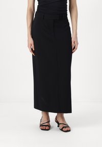 Jupe longue noire en tissu lisse, dotée d'une taille ajustée et d'une silhouette droite, associée à des sandales noires à lanières.