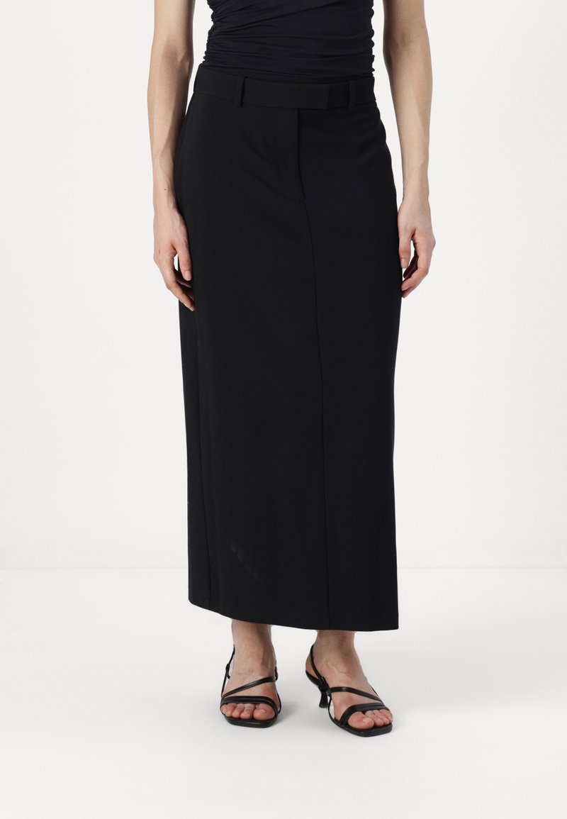 Jupe longue noire en tissu lisse, dotée d'une taille ajustée et d'une silhouette droite, associée à des sandales noires à lanières.