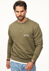 Olive green ribbed sweatshirt with crew neckline, light embroidery detail, and relaxed fit. Paired with light blue jeans and a watch.
