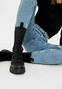 Černé semišové kotníkové boty s masivní podrážkou, hladkým povrchem, minimalistickým designem a poutkem na zadní straně. Nosené s modrými džíny.