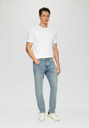 Man wearing white short-sleeve t-shirt, light blue jeans, and white sneakers standing with one hand in pocket against plain background.