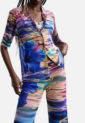 Žena oblečená v pestrofarebnom hodvábnom súprave košele a nohavíc s modrými ibištekom a abstraktnými vzormi odrazu vody, ruky v kapse.