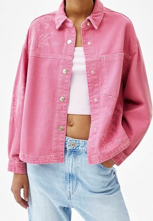 Une personne porte une veste rose boutonnée avec une broderie florale, un top court blanc et un jean bleu clair avec une main dans la poche.