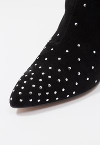 Schwarze Wildleder-Stiefelette mit einer spitzen Zehe, verziert mit verstreuten silbernen Nägeln auf der Außenseite für zusätzlichen Detail.