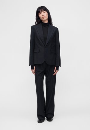 Vrouw staand, draag een zwarte op maat gemaakte blazer, een zwarte coltrui, broek met wijde pijpen en zwarte schoenen tegen een effen witte achtergrond.