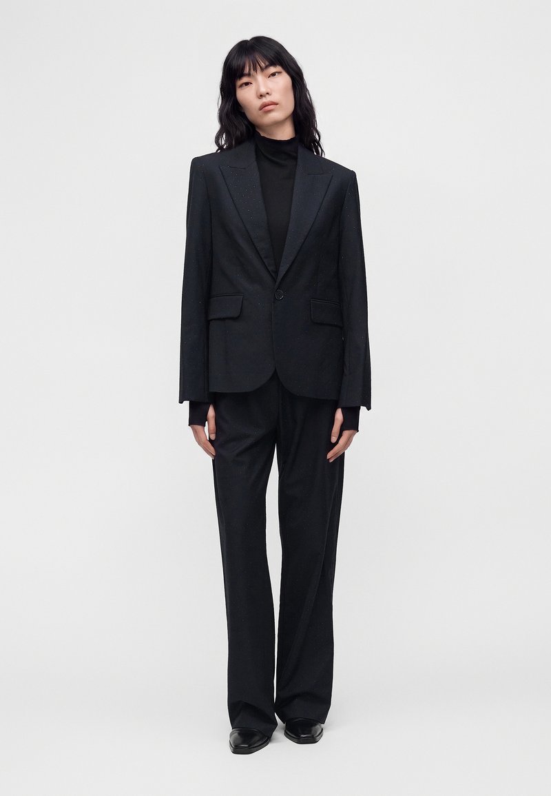 Γυναίκα που στέκεται, φοράει μαύρο tailored σακάκι, μαύρο ζιβάγκο, παντελόνια με φαρδιά μπατζάκια και μαύρα παπούτσια, μπροστά σε απλό λευκό φόντο.