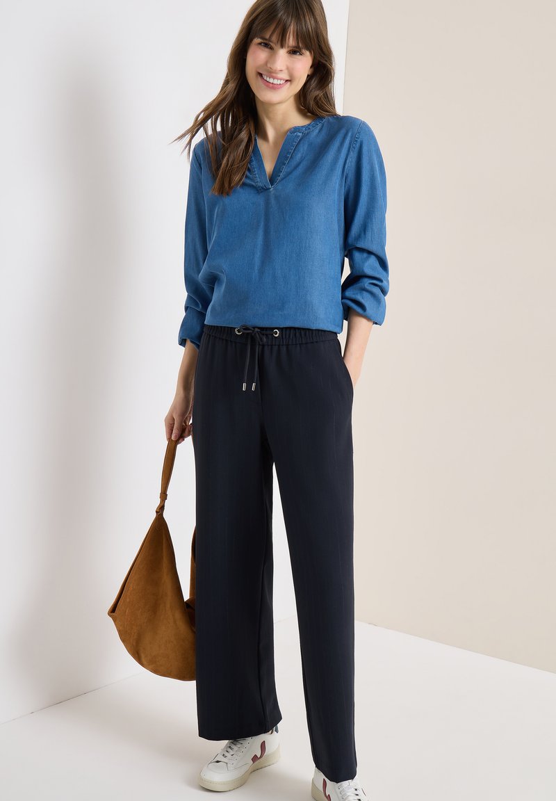 Femme souriante portant un chemisier bleu à manches longues, un pantalon noir à jambes larges, des baskets blanches, et tenant un sac à main marron sur un fond neutre.