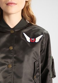 Svart satin bomberjacka med en dekorativ vinge-patch i röd och vit detalj, guldknappar och en dragkedjeficka på ärmen.