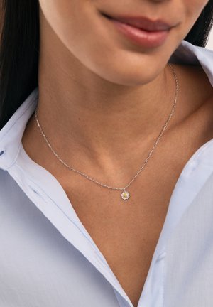 Close-up van een persoon die een fijne zilveren ketting met een kleine madeliefjessluiting draagt, gekleed in een lichtblauw overhemd.