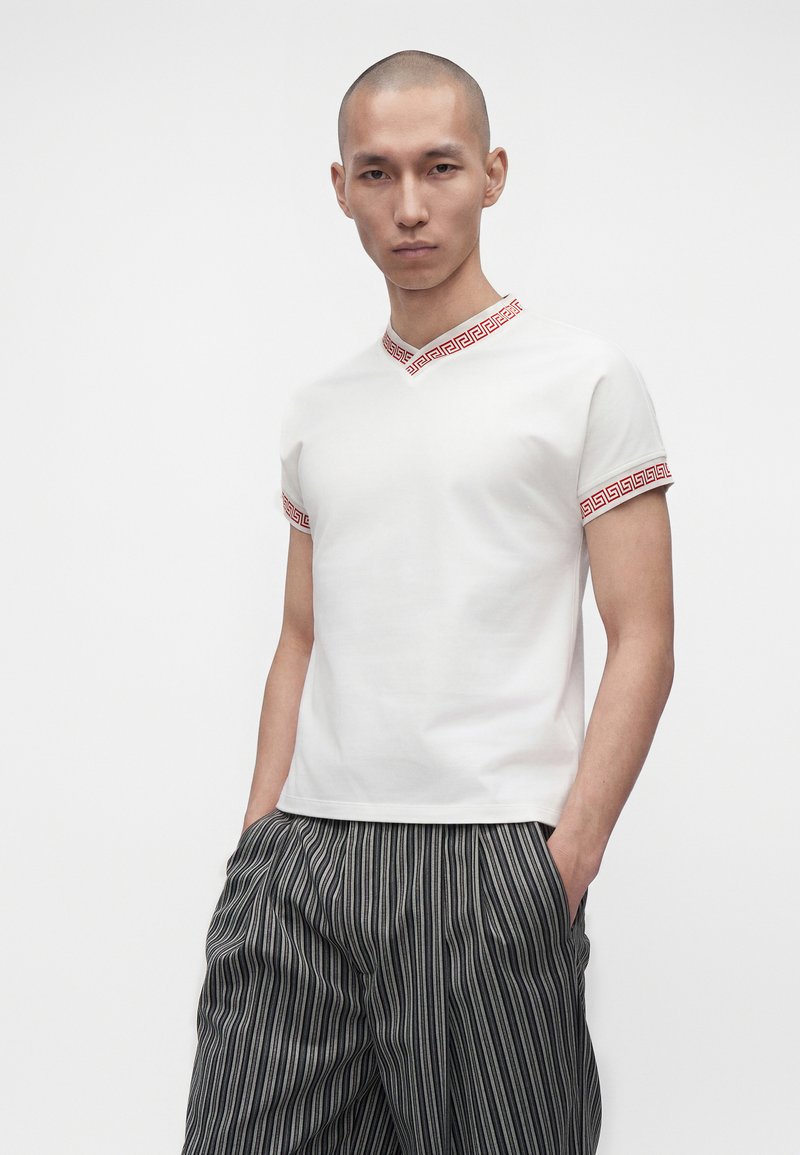 Homme aux cheveux courts portant une chemise blanche à manches courtes avec une bordure rouge à motif et un pantalon rayé noir et blanc, les mains dans les poches.