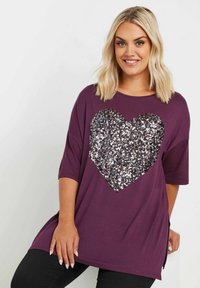 Camiseta oversized morada con un gran diseño de corazón de lentejuelas plateadas en la parte delantera, mangas cortas y aberturas en los laterales para facilitar el movimiento.