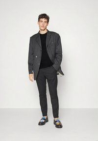 Blazer cinzento de abotoamento duplo sobre um suéter preto, calças cinzentas justas e sandálias plataforma coloridas com padrões artísticos.