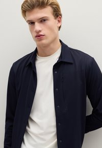 Camicia blu navy con bottoni e collo, caratterizzata da una texture liscia e una tasca sul petto. Indossata sopra una maglietta bianca a girocollo. Sfondo neutro.