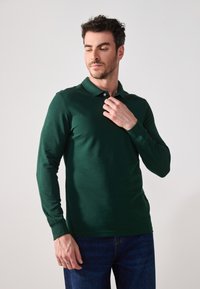 Grön långärmad polotröja med krage, med en knapp vid halsen, i kombination med denimjeans. Slät tygkvalitet och figurnära design.