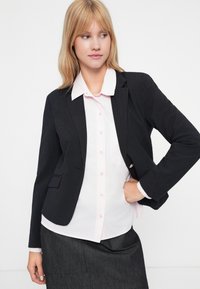 Blazer noir à rayures avec revers crantés, coupe slim et fermeture à un bouton sur une chemise rose pâle à boutons. Tissu texturé.