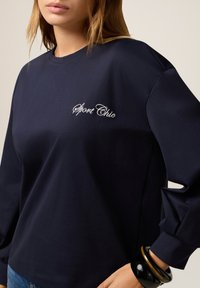 Donna che indossa una camicia a maniche lunghe blu navy con ricamo "Sport Chic" e bracciali neri e marroni al polso, su sfondo neutro.