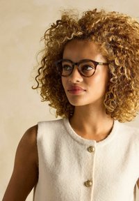 Hnedé okuliare s mušľovým vzorom sedia na vlnitých, kučeravých vlasoch. Krémový tielkový top má okrúhly výstrih a zlaté gombíky ako ozdobu.