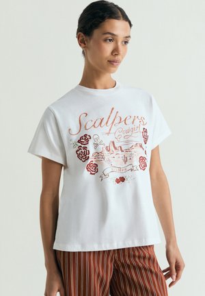 Joven mujer que lleva una camiseta blanca con estampado floral y de calaveras de estilo occidental y pantalones a rayas oxidadas, de pie frente a un fondo liso.