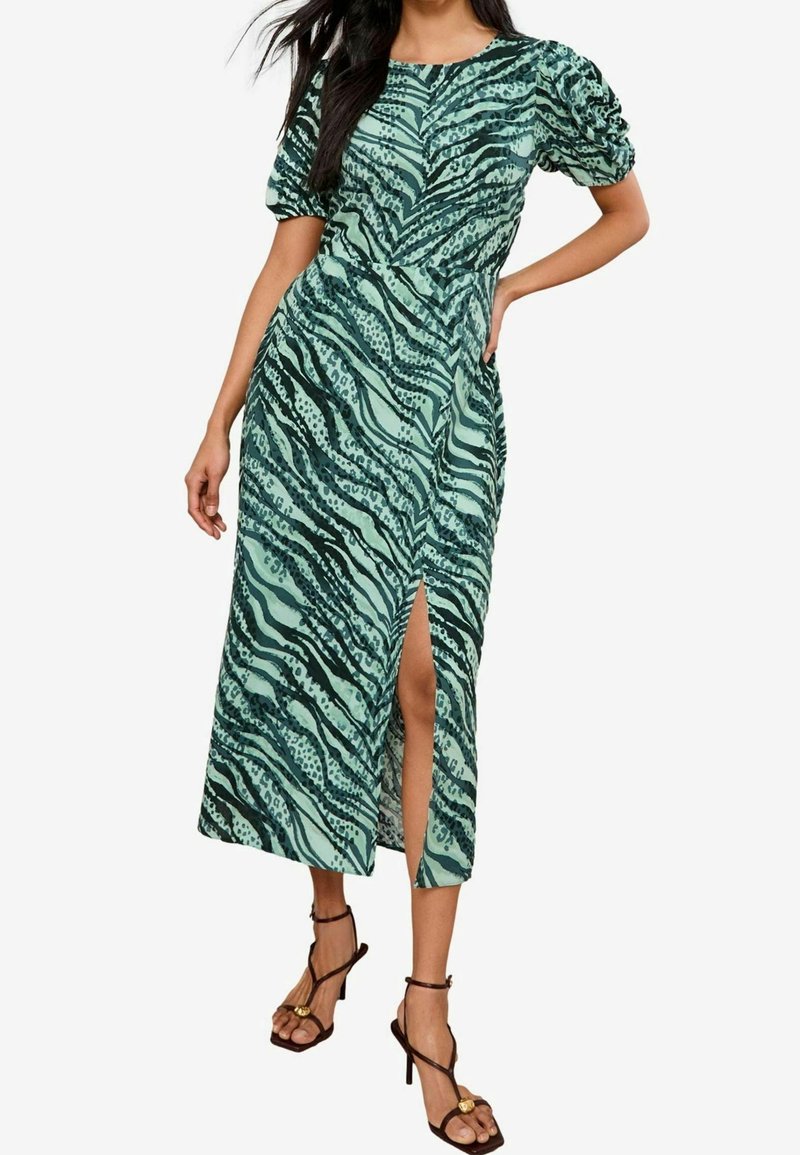 Femme portant une robe midi verte à imprimé animalier, manches courtes bouffantes, fente avant, et sandales noires à talons hauts avec brides à la cheville.