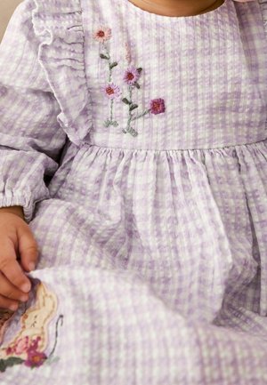 Kind, das ein lavendelfarbenes Karo-Kleid mit Rüschendetails trägt, bestickte Blumen auf der Brust und ein Hase, der in der Nähe des Saums gestickt ist.