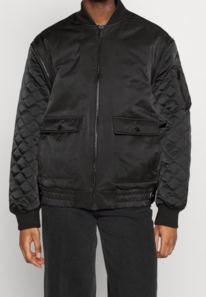 Personne portant une veste bomber matelassée noire avec fermeture éclair devant, poches à rabat, poche zippée sur le bras, et poignets et ourlet côtelés.