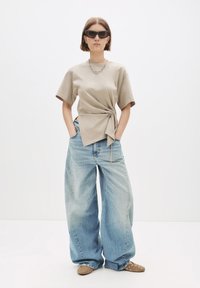 Kvinde iført oversized lyseblå jeans, beige indviklet t-shirt, brune naglede loafers, sorte solbriller og sølv kædedesmykker, stående med hænderne i lommerne.