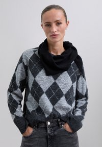 Pull en tricot argyle gris et noir avec un col roulé, featuring des poignets et un ourlet à côtes. Porté avec un jean noir sur un fond neutre.