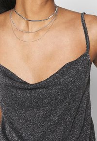 Svart ärmlös topp med en djup V-ringning, med en texturerad tyg med silverfläckar. Tre lager av silverhalsband hänger runt halsen.