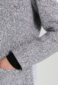 Šedý texturovaný svrchní oděv s měkkým, chlupatým povrchem; má otvor pro palec a ruku v přední kapse. Lehce a volně střižený design.