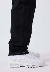 Persoon die zwarte broek en witte Nike Air Max sneakers draagt, staand op een lichte ondergrond tegen een effen achtergrond.