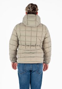 Giacca a vento trapuntata beige con cappuccio, caratterizzata da una superficie testurizzata e un motivo a griglia, abbinata a jeans blu.