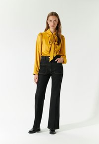 Blusa di raso gialla con dettaglio a fiocco, maniche lunghe. Jeans neri a vita alta con cuciture a contrasto e tasche posteriori decorative. Scarpe nere.