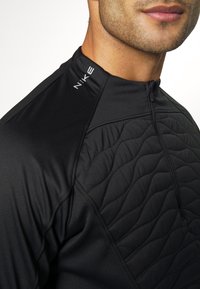 Gros plan sur le torse et le visage d'un homme portant une veste zippée noire Nike avec un panneau avant matelassé et un logo sur l'épaule.