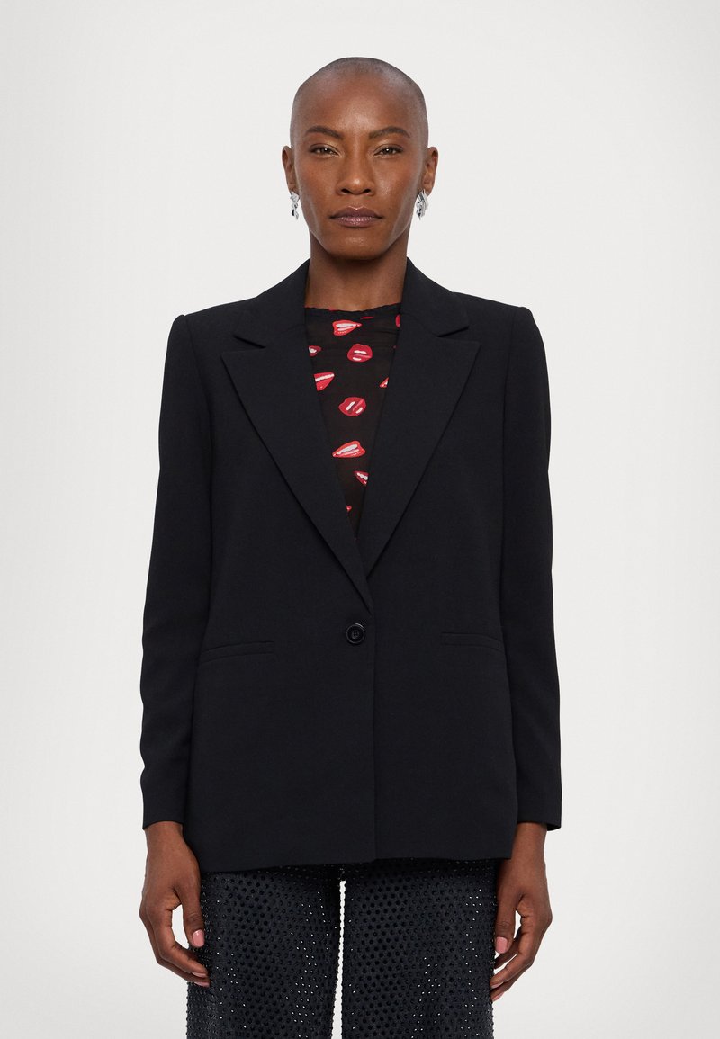Black tailored blazer with a single button, notch lapels, front pockets, and structured shoulders; worn over a patterned blouse and textured pants.