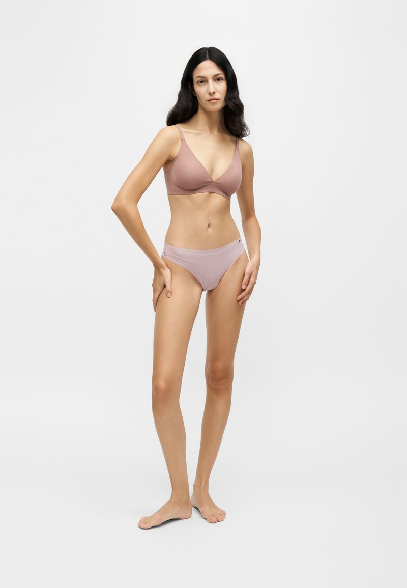 Femme aux longs cheveux foncés, debout pieds nus, portant un soutien-gorge mauve et des sous-vêtements assortis sur un fond blanc uni.