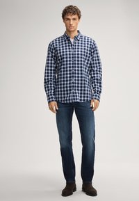Marineblaues und weißes karriertes Hemd mit langen Ärmeln, Knopfleiste und Ärmeldetails, kombiniert mit dunkelblauen Jeans und braunen Stiefeln.