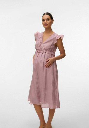 Gravid kvinde med mørkt hår sat tilbage, iført en lyserød ærmeløs graviditetskjole, stående med hænderne på maven.