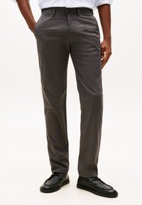 Homem a usar calças cinza-escuro de perna reta, sapatos pretos com atacadores e uma camisa branca enfiada para dentro com mangas arregaçadas.