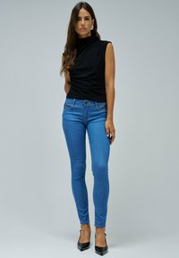 Mujer vestida con una blusa negra sin mangas, jeans ajustados azules, tacones negros puntiagudos y aretes de plata, de pie contra un fondo claro y liso.