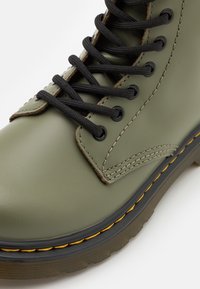 Bota de cuero verde oliva con cordones negros, punta redonda, costuras amarillas en contraste y suela de goma texturizada. Diseño clásico con ojales de metal.