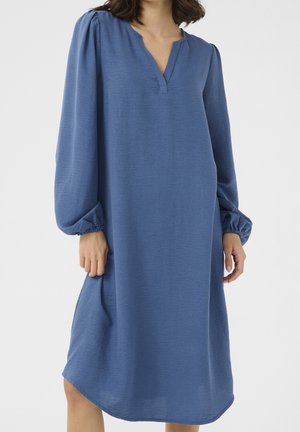 Femme portant une robe bleue fluide à manches longues avec un col en V et des poignets froncés, debout devant un fond uni.