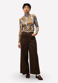 Langärmliges, eng anliegendes Oberteil mit einem braun-blau-weißen Muster, kombiniert mit hoch-taillierten, weit geschnittenen braunen Hose. Braune Schlappen.