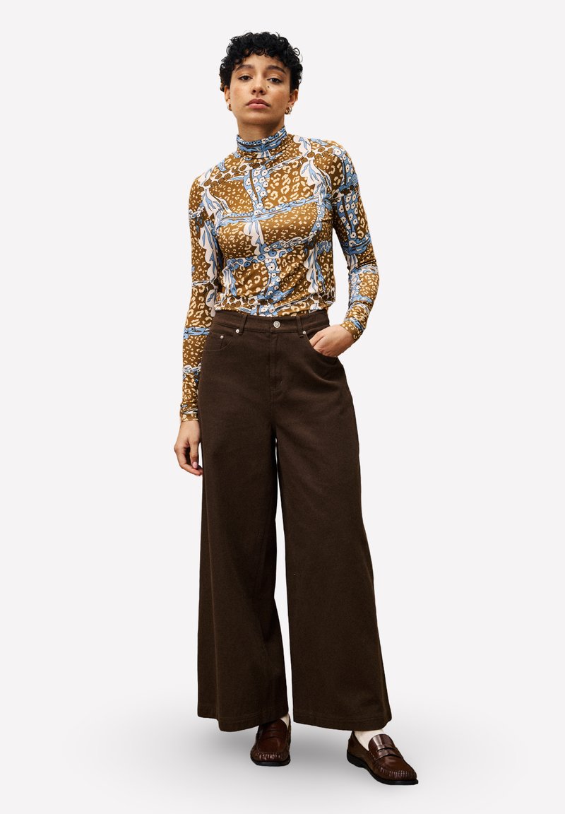 Langärmliges, eng anliegendes Oberteil mit einem braun-blau-weißen Muster, kombiniert mit hoch-taillierten, weit geschnittenen braunen Hose. Braune Schlappen.