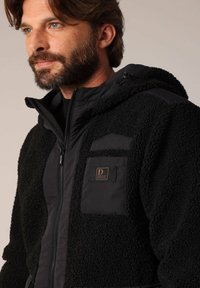 Zwarte fleecejack met een gestructureerde buitenlaag, nylon accenten, een ritssluiting aan de voorkant en een borstzak met een logo-opdruk.