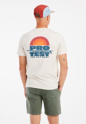 Vaaleanbeige t-paita, jossa on värikäs graafinen kuvasto, jossa on auringonlasku ja teksti "PRO TEST SUN SEA SURF", yhdistettynä vihreisiin shortseihin ja moniväriseen lippalakkiin.