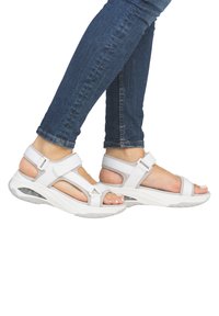 Sandales sportives blanches avec des sangles réglables, des semelles rembourrées et des accents texturés, associées à un jean bleu foncé.