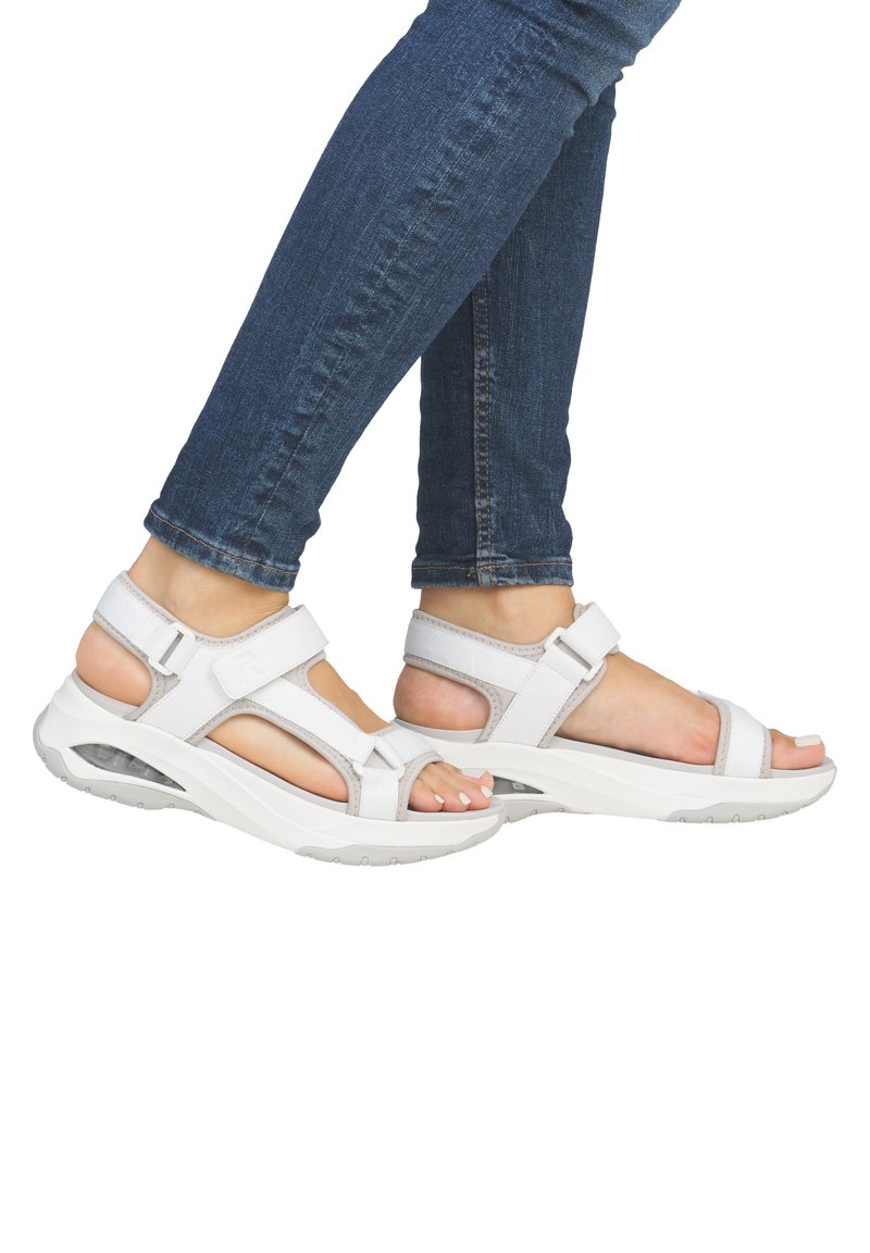 Sandales sportives blanches avec des sangles réglables, des semelles rembourrées et des accents texturés, associées à un jean bleu foncé.