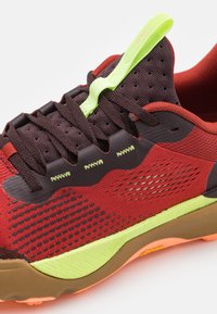 Rode en bruine sportschoen met een getextureerde mesh bovenkant, neon groene accenten en een rubberen zool. Voorzien van een geïntegreerd vetersysteem en ventilatiegaten.